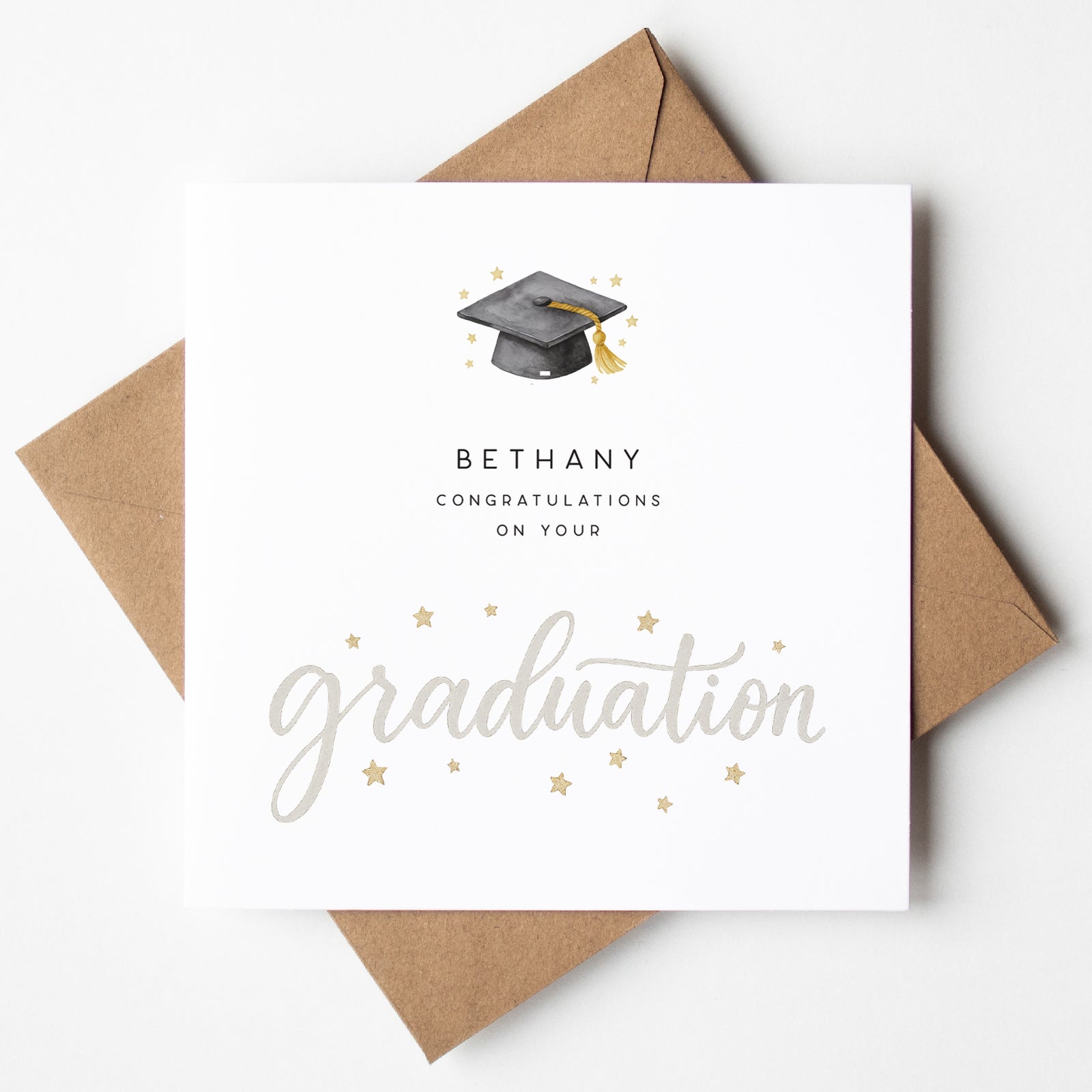 Graduation card with a black mortarboard and gold tassel above the name “Bethany,” featuring elegant grey script text reading “graduation” and scattered gold stars. Shown with kraft envelope.