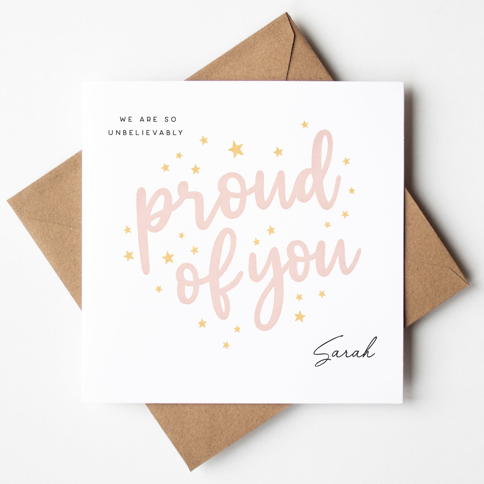 Personalised graduation card with blush pink script reading “Proud of You,” surrounded by scattered gold stars. Includes the name “Sarah” and a kraft envelope.