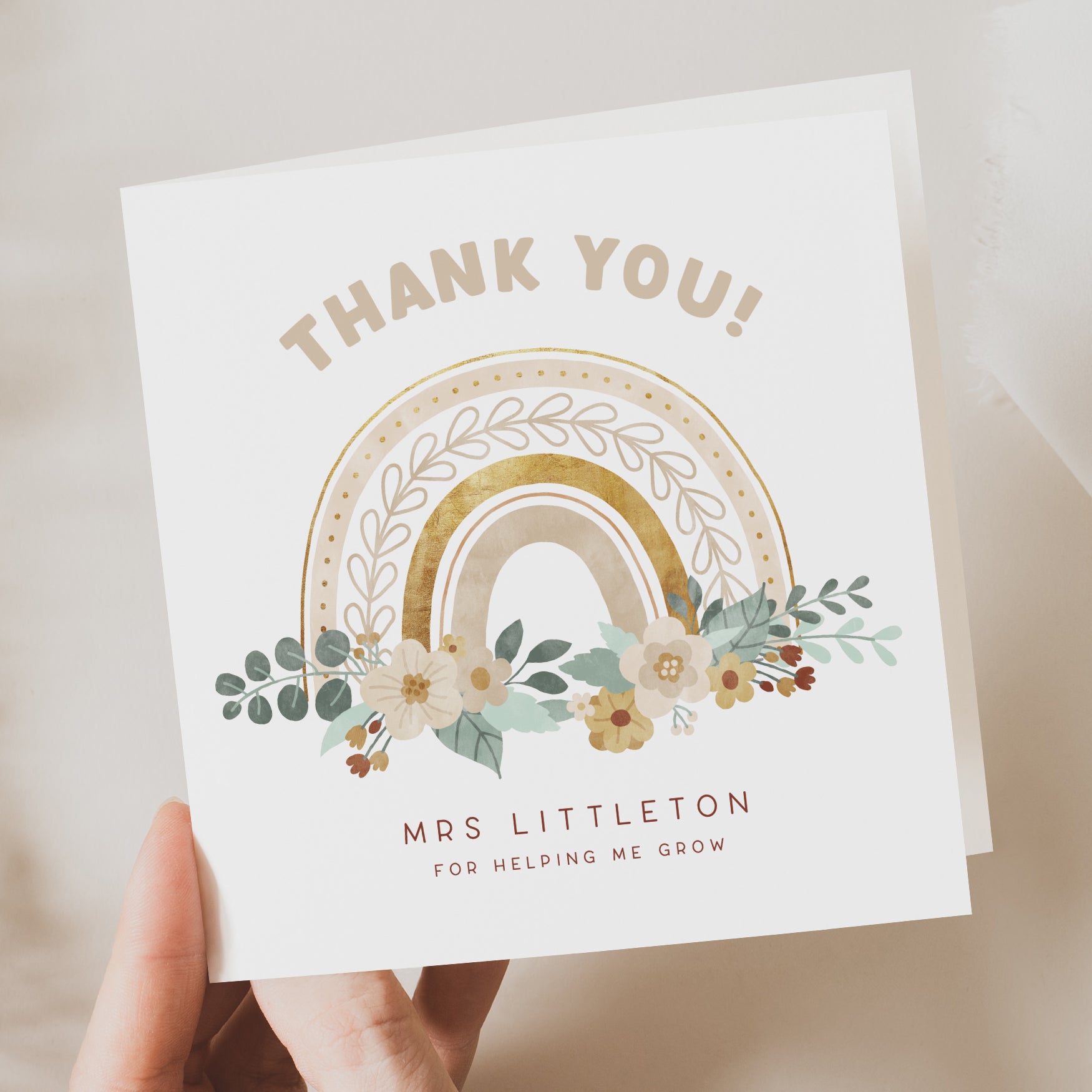 Hand holding a personalised teacher thank you card featuring a gold and beige floral rainbow illustration. Reads “Thank You!”, custom name line “Mrs Littleton – For helping me grow.”