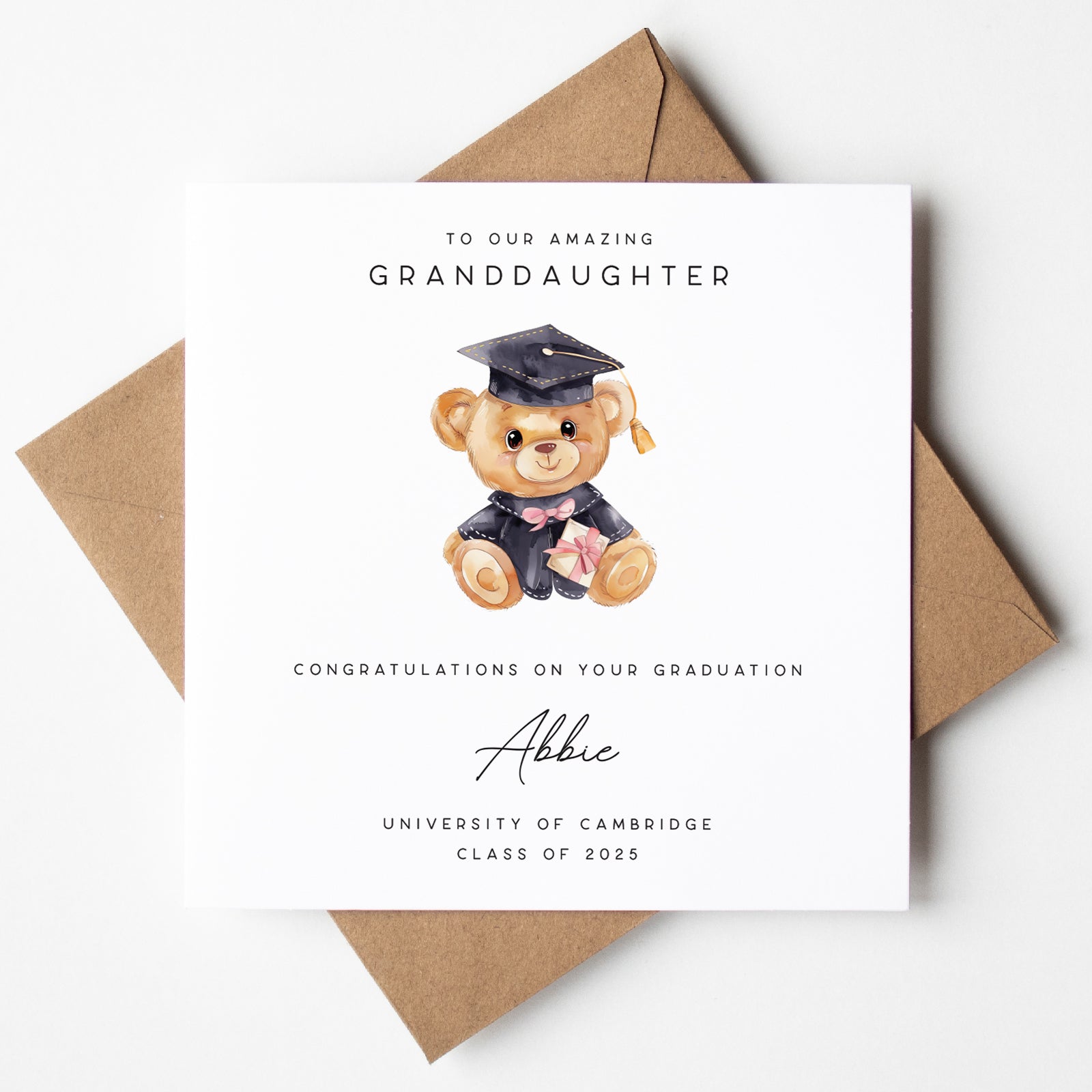 Personalised graduation card for a granddaughter, featuring a teddy bear in a graduation gown and cap holding a diploma with a pink ribbon. Custom text includes “To our amazing granddaughter,” the name “Abbie,” and “University of Cambridge – Class of 2025.” Card is paired with a kraft brown envelope.