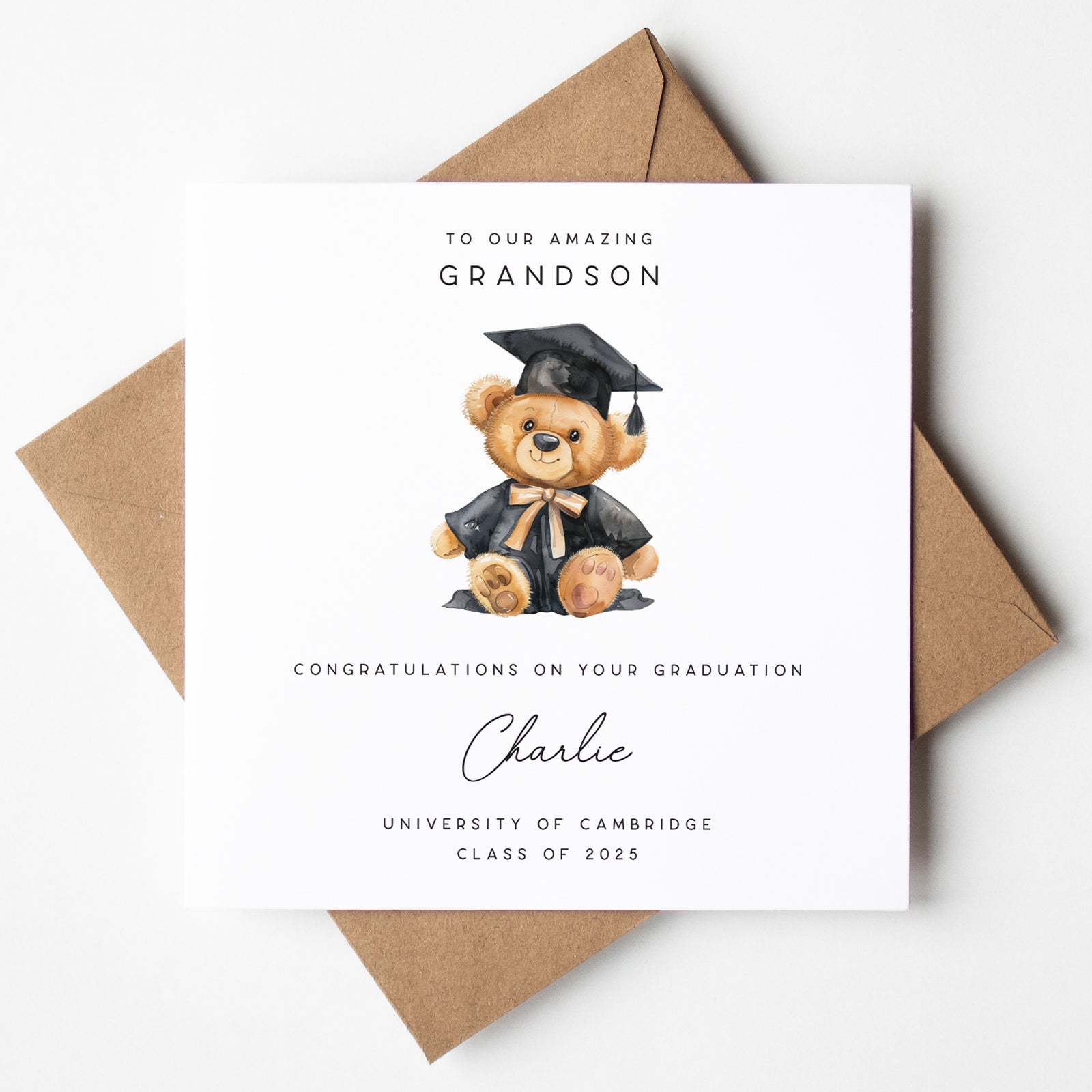 Personalised graduation card for a grandson, featuring a teddy bear in a black graduation gown and cap with gold trim. Includes the message “To our amazing grandson,” the name “Charlie,” and “University of Cambridge – Class of 2025.” Card is displayed with a kraft brown envelopE