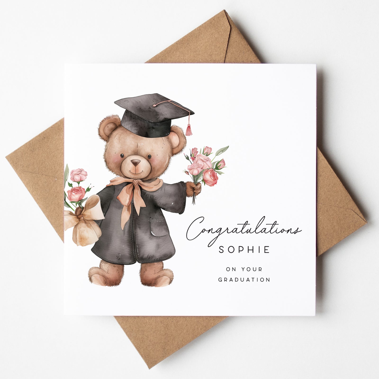Cute personalised graduation card featuring a smiling teddy bear in a graduation gown and cap, holding pink flower bouquets. Includes custom name “Sophie” and the message “Congratulations on your graduation.” Paired with a kraft brown envelope.