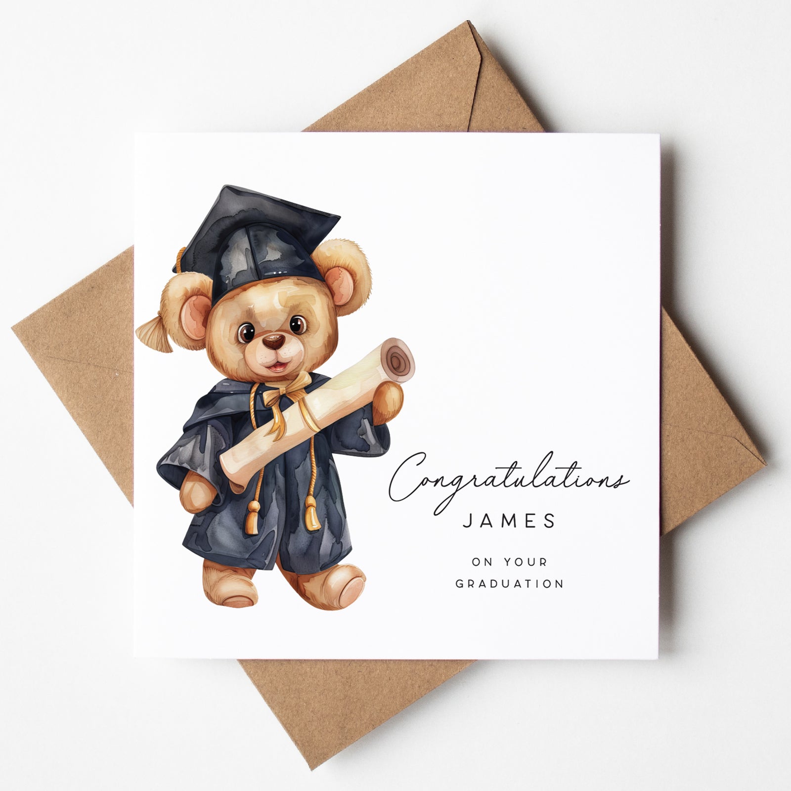 Personalised graduation card featuring a teddy bear in a black cap and gown, holding a graduation scroll tied with a gold ribbon. Includes custom name “James” with the message “Congratulations on your graduation.” Card is displayed with a kraft brown envelope.