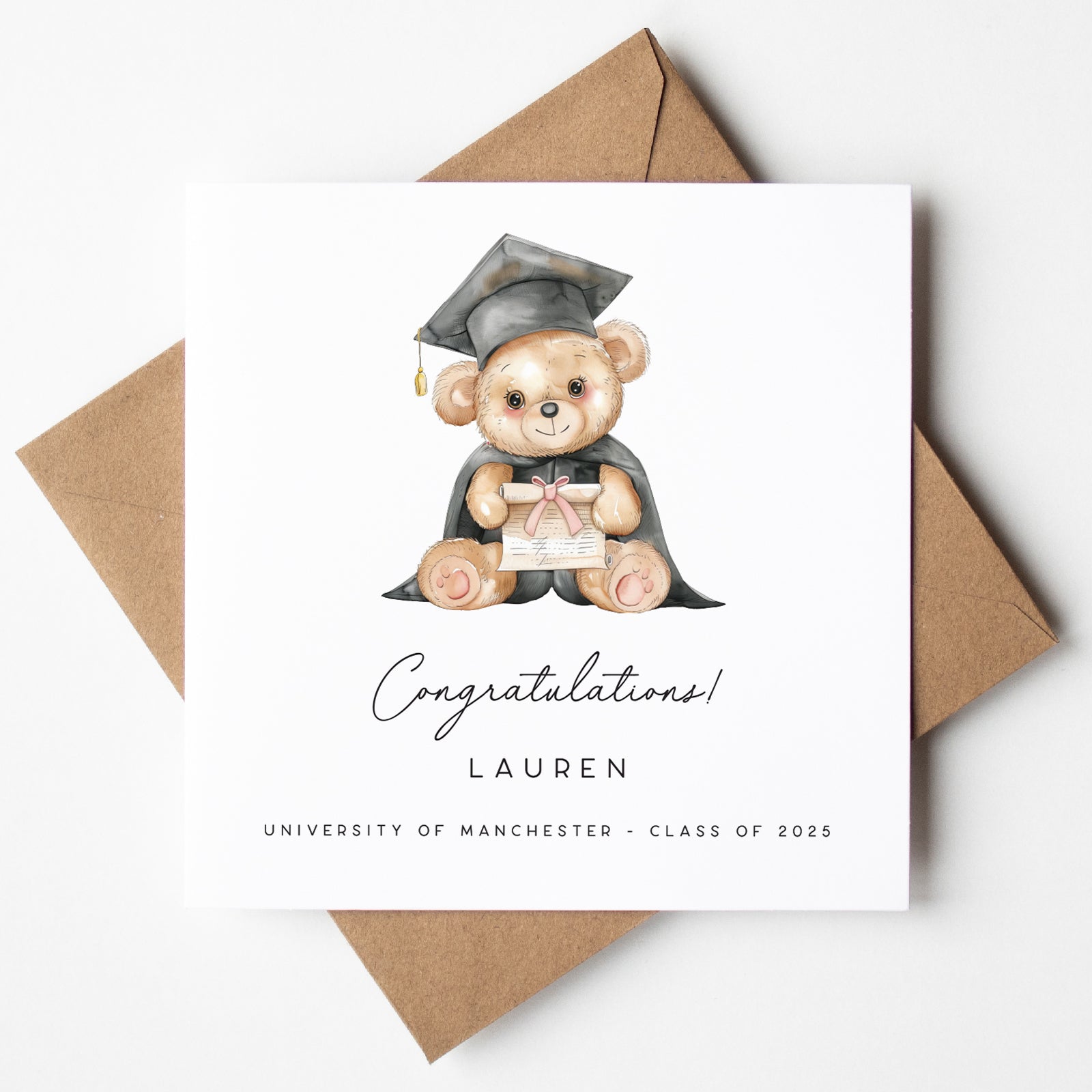 Personalised graduation card featuring a seated teddy bear in a cap and gown holding a diploma tied with a pink ribbon. Custom text includes “Congratulations,” the name “Lauren,” and “University of Manchester – Class of 2025.” Displayed with a kraft brown envelope.