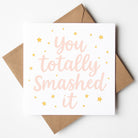 Pink and gold star-themed graduation card with the message “You totally smashed it” in soft script font. Displayed with a kraft brown envelope.